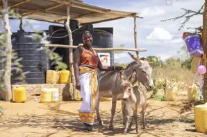 A woman stands with her donkey outside with a shelter and water barrels in the background