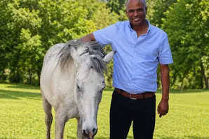 Yohannes Kassim stands outside, wearing a light blue shirt and black trousers, next to a white horse .
