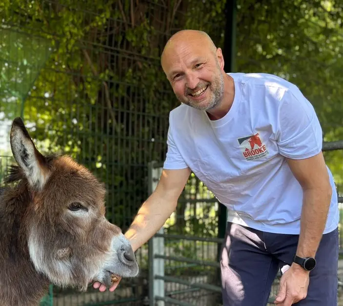 Brooke CEO Chris Wainwright wears blue trousers and a white t-shirt and smiles while petting a brown donkey