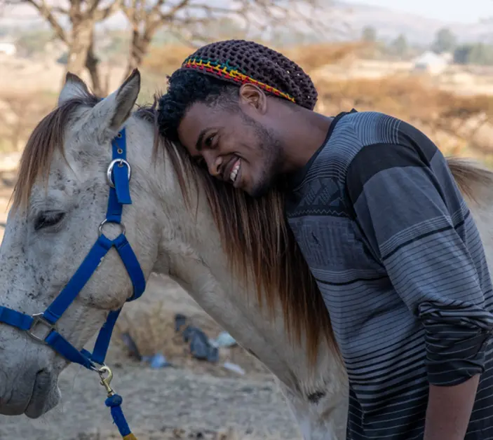 Tajebu, wearing a dark and light blue sweater, hugs the side of Bora, a white horse in blue harnessing.