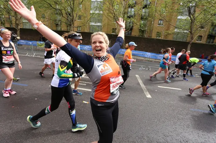 A person holds their arms up in joy and looks at the camera as they race a marathon with other runners in the background. They're wearing a grey, orange and black Brooke top and dark paints.