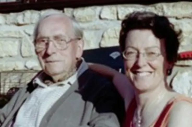 Diana, wearing a pink blouse, sits with her dad, dressed in a white shirt and grey cardigan, outside a house.