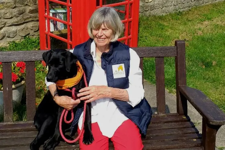 Sandi Cole, a Brooke supporter, sits on a bench with her black labrador Molly, in front of a red telephone box.